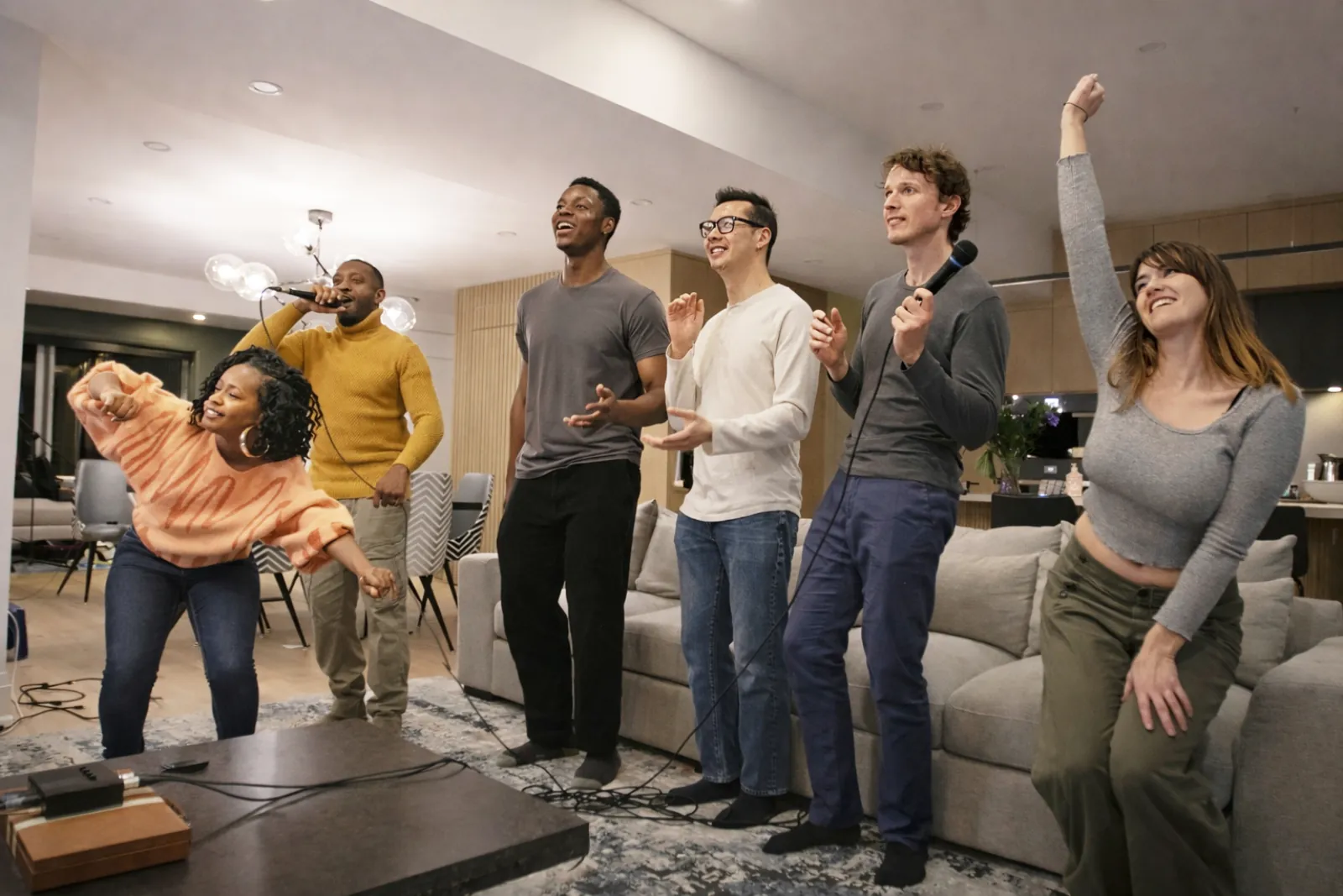 Six friends standing and singing together in a living room, two of them holding microphones